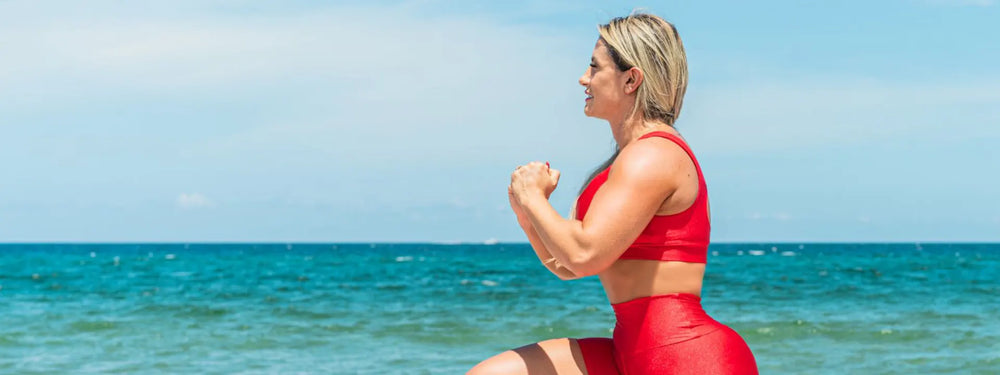Woman in a red outfit exercising on a beach with clear blue water and sky.
